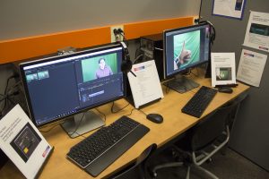Two desktop computers on a bench. The image of a person in front of a green screen is visible on one of the computer screens.