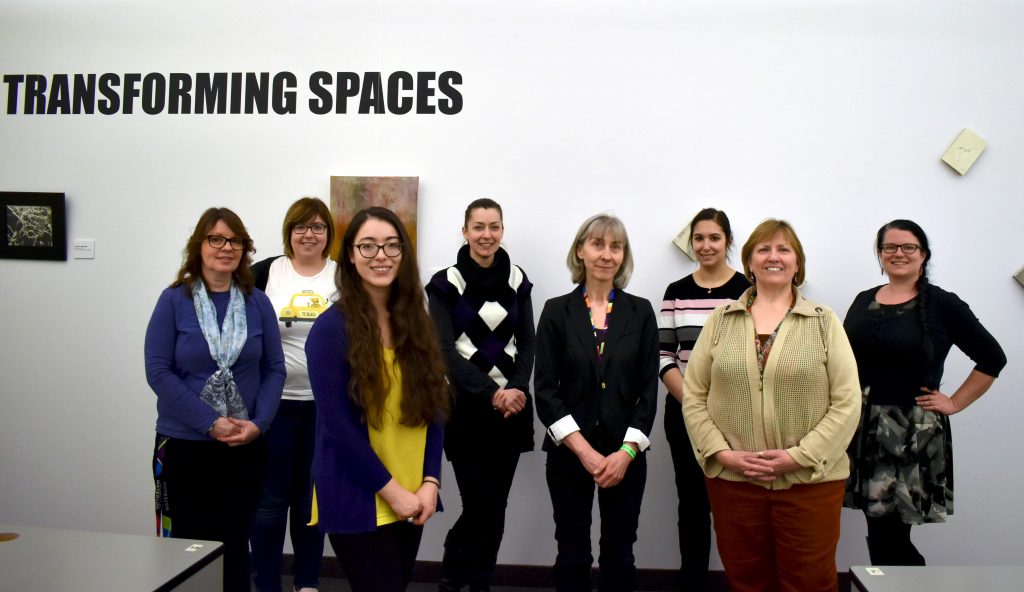 The image represents a group of 8 women standing in front of the exhibition wall. The words Transforming Spaces are printed on the wall. Art can be partly seen behind the group.