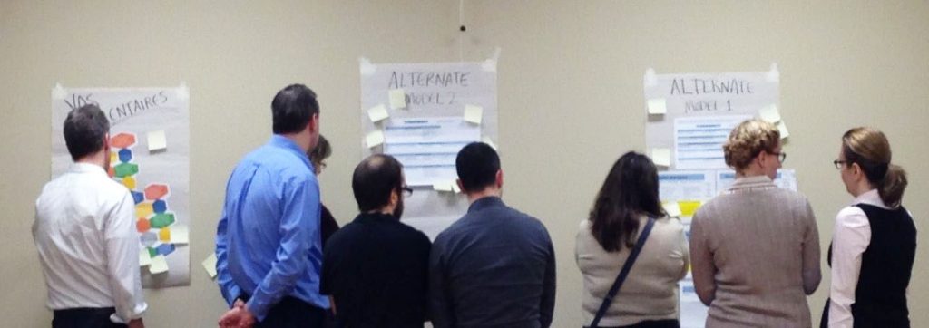 A group of people, their back to the camera, are observing sheets of paper and sticky notes taped to a wall.