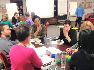 People engaged in discussion around a table, on which there are colourful play blocks, sheets of paper and sticky notes. Another table is seen in the background, as well as a person standing reading a flip chart (illegible text).