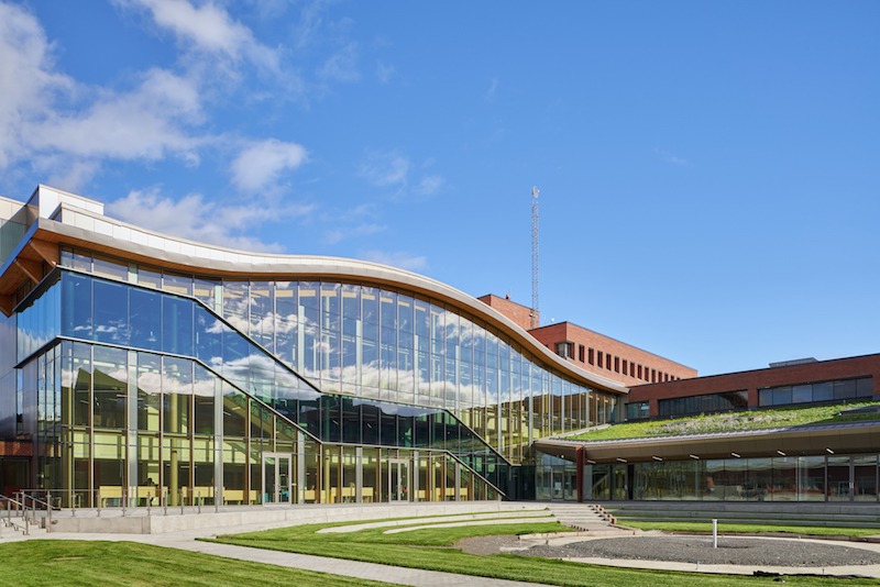 Exterior view of the new DARE District building at Algonquin College. It is a modern building with a glass facade that reflects the blue sky with a few clouds.