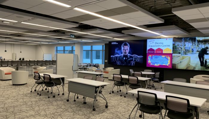 Image Of The Digital Creativity Centre At Mohawk College. A Row Of Tables Faces A Large Screen.