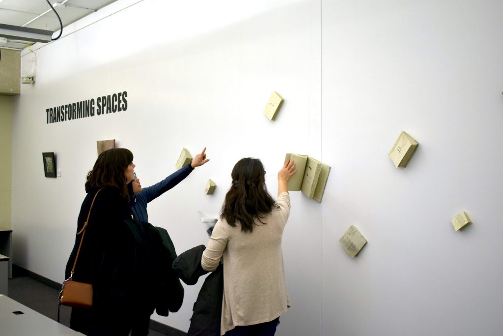 Three women, their back to the camera, are seen interacting with an art installation involving books attached to a wall. One is flipping through the pages of one of the attached books. The words Transforming Spaces are printed on the wall.