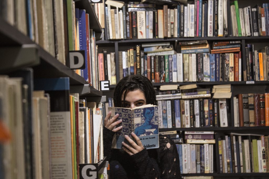 Photo of a woman surrounded by books, covering her face with a copy of 1984.