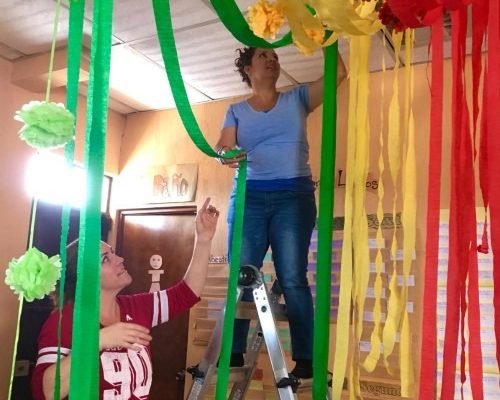 Two Women, One Standing On A Ladder, Hanging Red, Yellow, Blue And Green Streamers