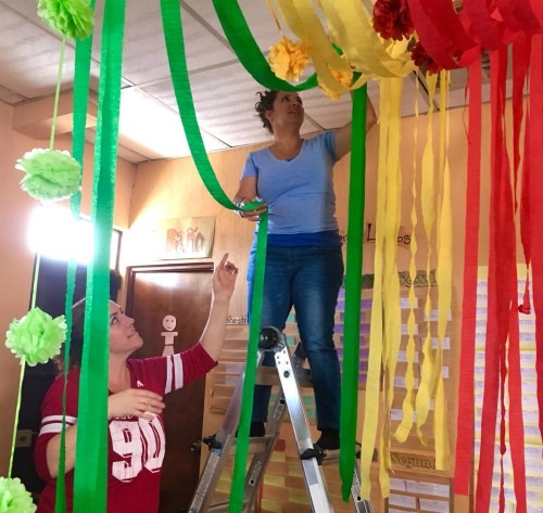 Two women, one standing on a ladder, hanging red, yellow, blue and green streamers