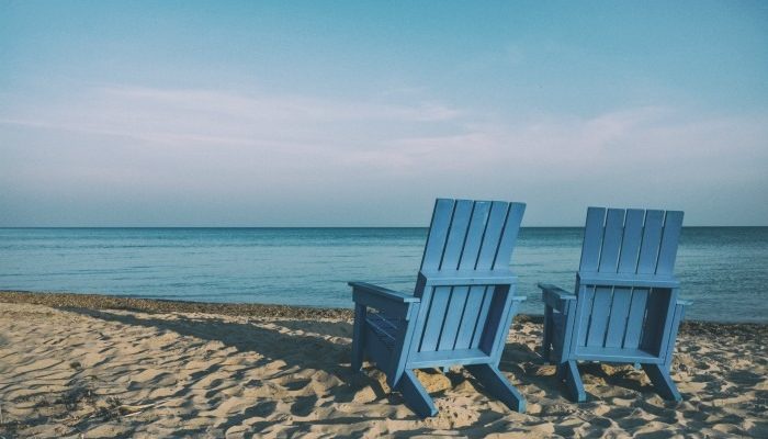 Two Blue Adirondack Chairs On A Beach Facing The Ocean