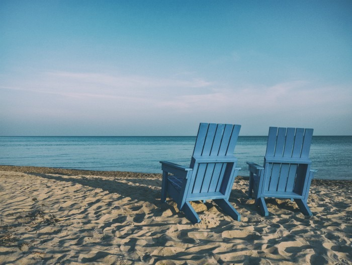 Two blue adirondack chairs on a beach facing the ocean