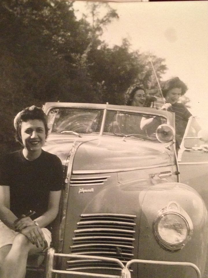 A photo of a girl sitting on the from bumper of an old car with two girls to her left in the back.