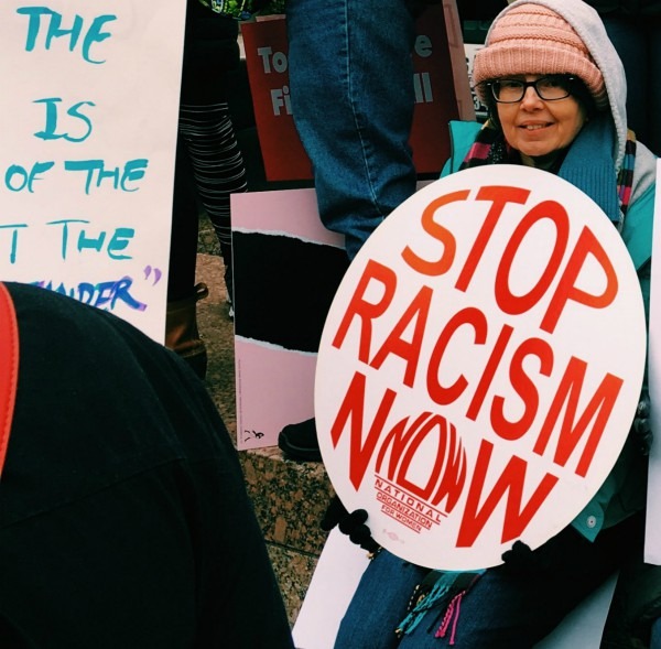 A woman sitting holding a sign that says Stop Racism Now