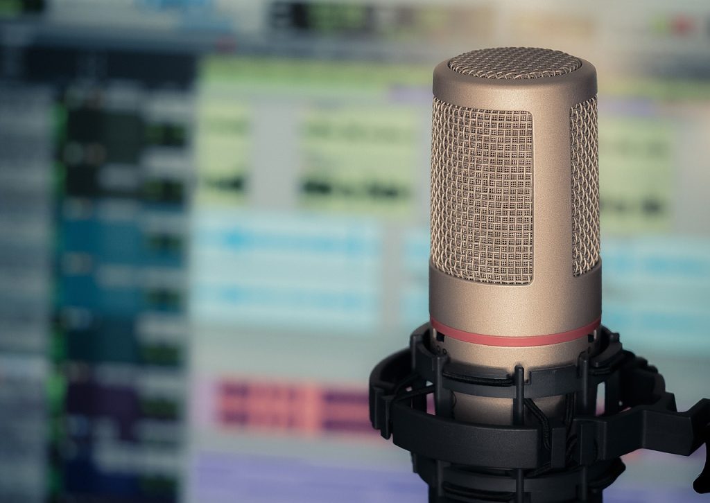 Picture of a microphone in the foreground and a blurry computer screen with audio editing software in the background