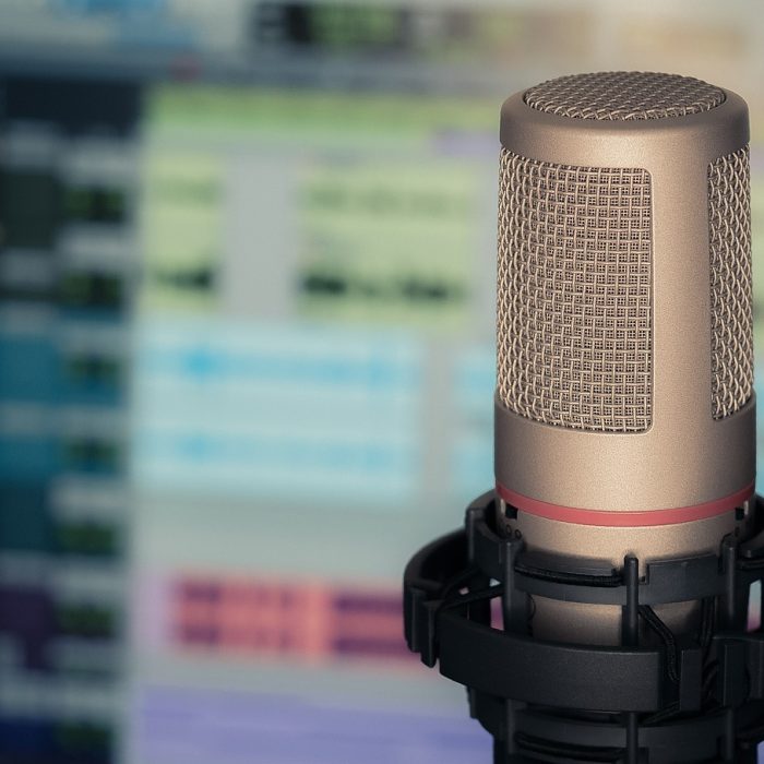 Picture Of A Microphone In The Foreground And A Blurry Computer Screen With Audio Editing Software In The Background